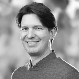 A person with short hair smiles in a black and white portrait, wearing a sweater with a blurred background.