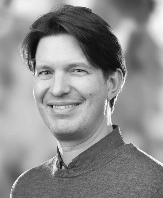 A person with short hair smiles in a black and white portrait, wearing a sweater with a blurred background.